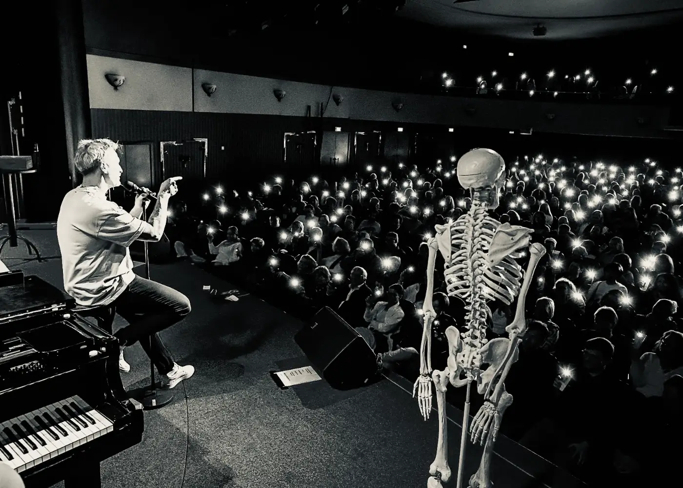 der-physiopath-live-show-bühne-entertainer-physiotherapie Der Physiopath sitzt auf der Bühne mit Mikrofon neben einem Skelettmodell, Publikum hält Handys mit Licht in die Höhe.